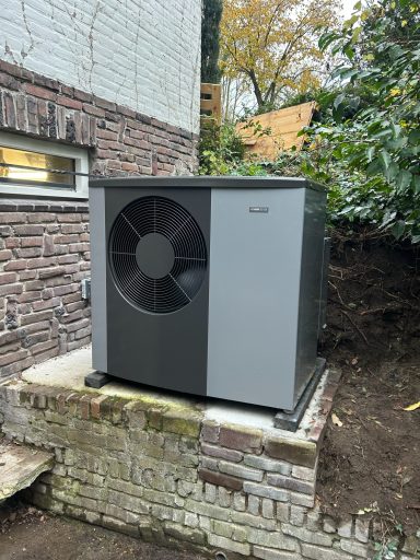 Buitenunit van een warmtepomp naast een bakstenen muur en groenere omgeving.